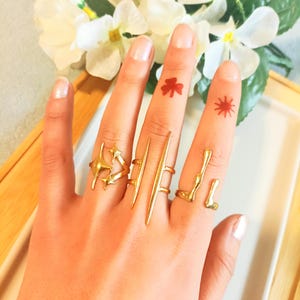 Könnte beinhalten: Eine Hand mit mehreren goldfarbenen Ringen in verschiedenen Designs, darunter Stern- und lineare Formen. Die Finger weisen auch kleine rote Tätowierungen auf, ein Kleeblatt und ein Sonnenstrahl.