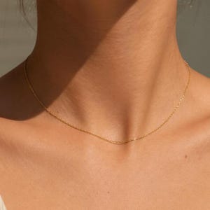 Pode incluir: Um delicado colar de corrente de ouro com um design de elos finos e delgados que repousa sobre o pescoço. O metal tem um acabamento polido e reflexivo que capta a luz, criando uma aparência sutil e discreta para esta peça de joalharia.