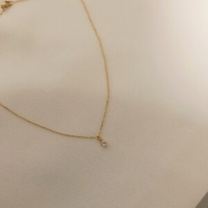 May include: A delicate gold chain necklace featuring a single, small, clear round-cut gemstone pendant suspended from the center. The fine metal links form a simple, slender chain that secures with a small clasp visible at the top corner.