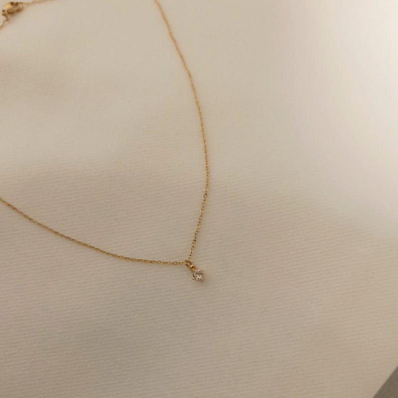 May include: A delicate gold chain necklace featuring a single, small, clear round-cut gemstone pendant suspended from the center. The fine metal links form a simple, slender chain that secures with a small clasp visible at the top corner.