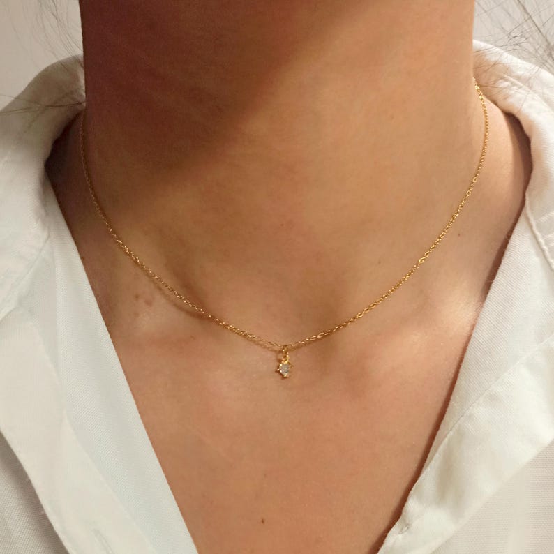 May include: A delicate gold chain necklace featuring a single, small, clear, round-cut gemstone pendant centered on the chain. The necklace rests against the neck, showcasing a simple, refined design with a polished metallic finish and a secure clasp.