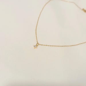 May include: A delicate gold chain necklace featuring a single, small, clear, round-cut gemstone pendant suspended from the center. The fine metal links form a simple, continuous chain that curves gracefully across the frame, highlighting the solitaire stone.