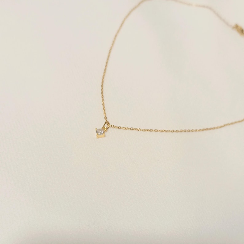 May include: A delicate gold chain necklace featuring a single, small, clear, round-cut gemstone pendant suspended from the center. The fine metal links form a simple, continuous chain that curves gracefully across the frame, highlighting the solitaire stone.
