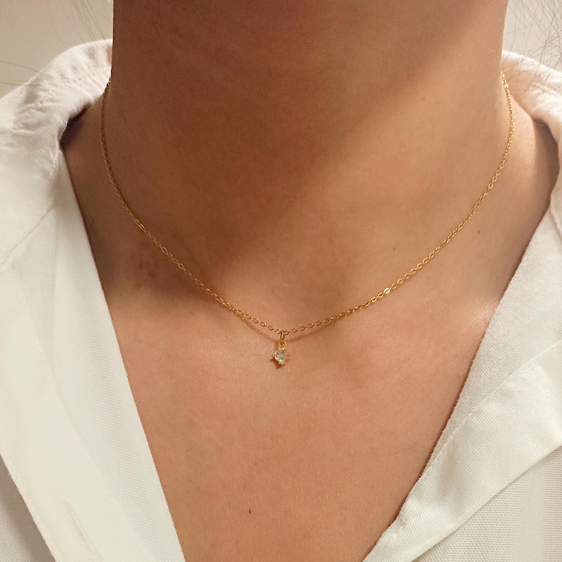 May include: A delicate gold-tone chain necklace featuring a single, small, clear round-cut gemstone pendant centered on the chain. The necklace rests against the neck, showcasing a simple link design with a secure clasp-free appearance.
