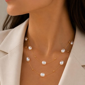Può includere: Una collana a doppia catena dorata con perle barocche bianche e piccole sfere dorate distanziate uniformemente. Le sottili catene in metallo sono disposte su due livelli, alternando perle e sfere lungo tutta la lunghezza dei collegamenti.