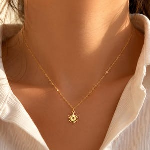 Può includere: Una collana color oro caratterizzata da una catenina sottile e un piccolo ciondolo piatto a forma di sole raggiante. Il ciondolo è centrato sulla catena che poggia sul collo. Il metallo ha una finitura lucida che riflette la luce.