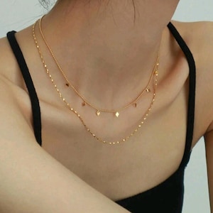 Könnte beinhalten: Eine goldfarbene Halskette mit zwei Strängen. Der obere Strang hat kleine rautenförmige Anhänger, während der untere Strang ein filigranes Kettenmuster aufweist. Die Halskette wird über einem schwarzen Oberteil getragen.