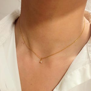 May include: A delicate gold chain necklace featuring a single, small, clear, four-pronged solitaire gemstone pendant centered on the collarbone. The thin, polished gold-tone metal chain rests against the skin, highlighting the minimalist design.
