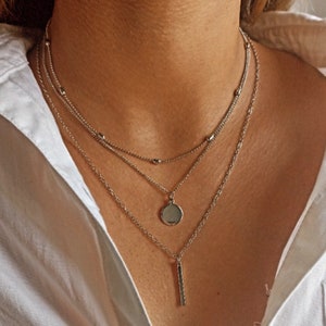 Peut inclure: Collier argenté à plusieurs rangs. La chaîne supérieure est ornée de petites perles, la chaîne du milieu est délicate, et la chaîne inférieure comporte un pendentif rond et une barre verticale. Le collier est porté sur une chemise blanche.