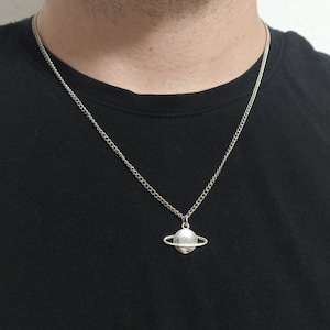 Könnte beinhalten: Silberfarbene Halskette mit einem Planetenanhänger. Der Anhänger hat eine strukturierte Oberfläche und einen Ring, der an einer filigranen Kette hängt. Die Halskette wird auf einem schwarzen T-Shirt getragen.