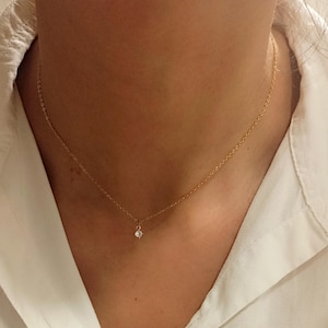 May include: A delicate gold chain necklace featuring a single, small, round, clear gemstone pendant centered at the base of the neck. The thin metal chain rests against the skin, highlighting the minimalist design of the solitaire stone setting.