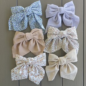Könnte beinhalten: Eine Auswahl von sechs großen Stoff-Haarschleifen in verschiedenen Farben und Mustern. Die Schleifen umfassen Blumenmuster in Blau, Beige und Weiß sowie unifarbene Varianten wie Lavendel und Beige. Die Schleifen sind auf einer neutralgrauen Oberfläche angeordnet.