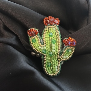 Könnte beinhalten: Ein bestickter Kaktus-Broschen-Anstecker mit einem grünen Körper, roten Blütenakzenten und goldenen und grünen Perlendetails. Die Brosche ist vor einem schwarzen Hintergrund platziert, der die aufwendige Perlenarbeit zeigt.