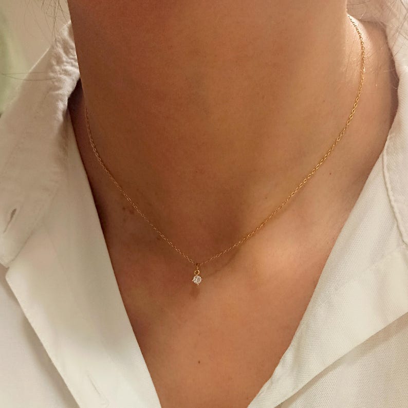 May include: A delicate gold chain necklace featuring a single, small, round, clear gemstone pendant centered on the chain. The necklace rests against the neck, showcasing a thin, polished metal link design with a secure, minimalist setting.