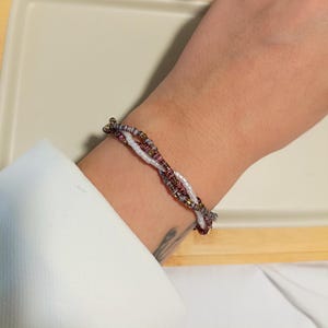 Può includere: Un braccialetto di perline con un design intrecciato. Il braccialetto presenta piccole perline colorate nelle tonalità del bianco, del viola e dell'oro. Il braccialetto è indossato su un polso e lo sfondo è di colore neutro.