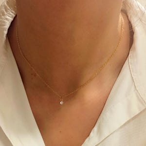 May include: A delicate gold chain necklace featuring a single, small, clear round-cut gemstone pendant centered at the base. The necklace is worn around the neck, showcasing a thin, polished metal link chain and a simple, secure prong setting.