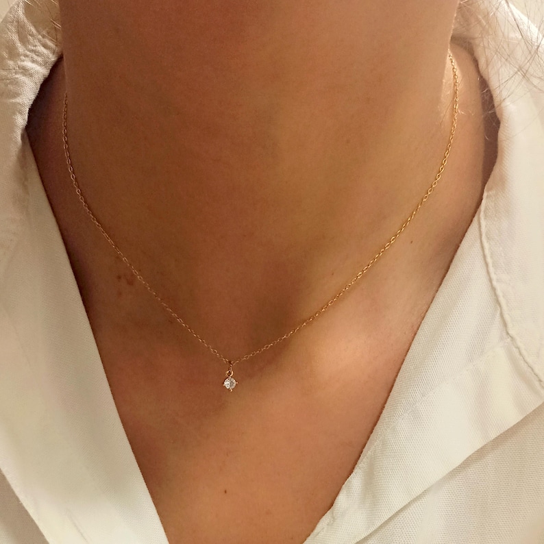 May include: A delicate gold chain necklace featuring a single, small, clear round-cut gemstone pendant centered at the base. The necklace is worn around the neck, showcasing a thin, polished metal link chain and a simple, secure prong setting.