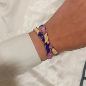 Könnte beinhalten: Zwei Armbänder mit einem geometrischen Muster aus blauen, rosa- und cremefarbenen Perlen. Die Armbänder werden am Handgelenk getragen, und die Perlen sind auf einem kupferfarbenen Draht aufgefädelt.