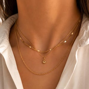 Puede incluir: Un conjunto de collares dorados en capas que consta de tres cadenas distintas. La cadena superior presenta eslabones metálicos planos, la cadena central sostiene un pequeño colgante redondo con una piedra clara engastada y la cadena inferior es una cadena de eslabones sencilla y delicada.