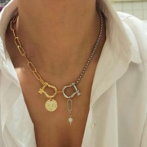 Può includere: Una collana bicolore con una catena dorata a sinistra e una catena argentata a destra. Il lato dorato presenta un ciondolo circolare con un motivo a occhio, mentre il lato argentato ha un piccolo pendente a stella. La collana è indossata su una camicia bianca.