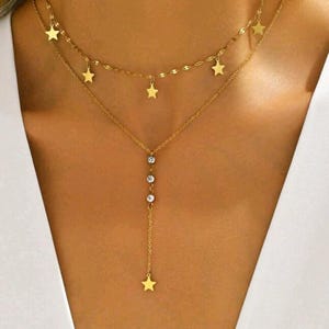 Può includere: Una collana dorata a più strati con dettagli a stella e cristallo. Lo strato superiore presenta piccoli ciondoli a stella, mentre lo strato inferiore ha una catena verticale con tre cristalli e un singolo pendente a stella. La collana è indossata su uno sfondo bianco.