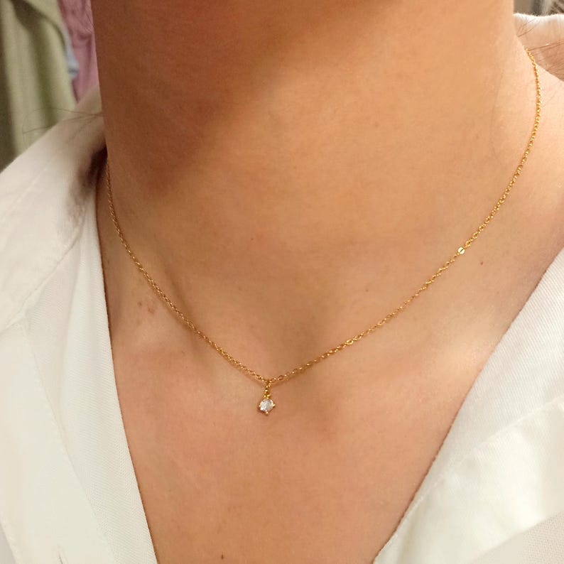 May include: A delicate gold chain necklace featuring a single small, clear, round-cut gemstone pendant centered on the chain. The necklace rests against the neck, showcasing a thin, polished metal link design with a secure clasp connection.