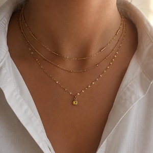 Könnte beinhalten: Ein dreilagiges Goldhalskettenset mit drei verschiedenen Ketten. Die oberste Kette hat kleine Goldstäbchen, die mittlere Kette zarte Perlen und die unterste Kette einen kleinen, runden, gelben Edelsteinanhänger in einer Goldfassung.