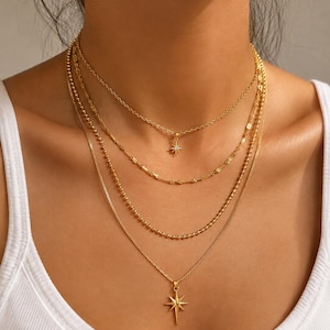 May include: A layered gold-tone necklace set consisting of four distinct chains. The top chain features a small starburst pendant with a central stone, followed by a textured chain, a beaded chain, and a bottom chain with a larger starburst pendant.
