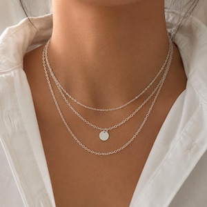 Könnte beinhalten: Eine mehrlagige silberfarbene Halskette bestehend aus drei zarten Ketten unterschiedlicher Länge. Die mittlere Kette ist mit einem kleinen, glatten, kreisförmigen Anhänger versehen. Die Ketten bestehen aus feinen, ovalen Gliedern.