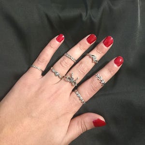 Könnte beinhalten: Eine Hand mit mehreren silbernen Ringen in verschiedenen Designs, darunter ein verdrehtes Band, ein kettenartiger Ring, ein Knoten-Design, ein Ring mit floralem Design und ein Ring mit kleinen Steinen. Die Nägel sind rot lackiert.