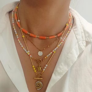 Könnte beinhalten: Ein mehrlagiges Halsketten-Set mit verschiedenen Perlen und Anhängern. Die oberste Schicht hat orangefarbene rechteckige Perlen, während andere kleine weiße, gelbe und goldene Perlen enthalten. Ein Strang hat einen kleinen goldenen Schildkrötenanhänger und ein weiterer einen Muschelanhänger.