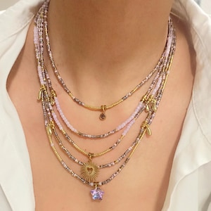 May include: A multi-strand necklace with small, colourful beads in shades of pink, gold, and grey. The necklace includes gold-toned charms and pendants. One pendant is a star-shaped charm with a purple crystal, and another is a sunburst design.