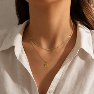 Könnte beinhalten: Eine goldfarbene, mehrlagige Halskette mit zwei zarten Ketten. Die kürzere Kette ist mit kleinen zylindrischen Metallperlen verziert, während die längere Kette einen kleinen, flachen Anhänger in Form einer achtblättrigen Blume trägt.