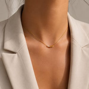 Könnte beinhalten: Eine zarte Goldkettenhalskette mit einer kleinen Ansammlung von fünf winzigen, polierten Goldperlen in der Mitte. Die Halskette liegt am Hals an und zeigt ein schlichtes, raffiniertes Design mit einer dünnen Metallgliederkette und einem warmen Glanz.