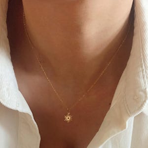 Pode incluir: Um delicado colar dourado com um pingente de sol. O sol, com seus raios, pende de uma corrente fina, criando um visual minimalista e elegante. O colar é usado sobre uma camisa branca.