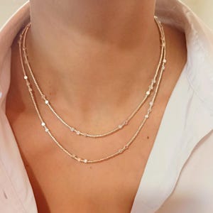 Op de afbeelding: Een delicate, gelaagde ketting met twee strengen kleine, ronde kralen en afgewisseld met kleine, platte, cirkelvormige bedels. De ketting is lichtgoudkleurig en rust op een wit overhemd.