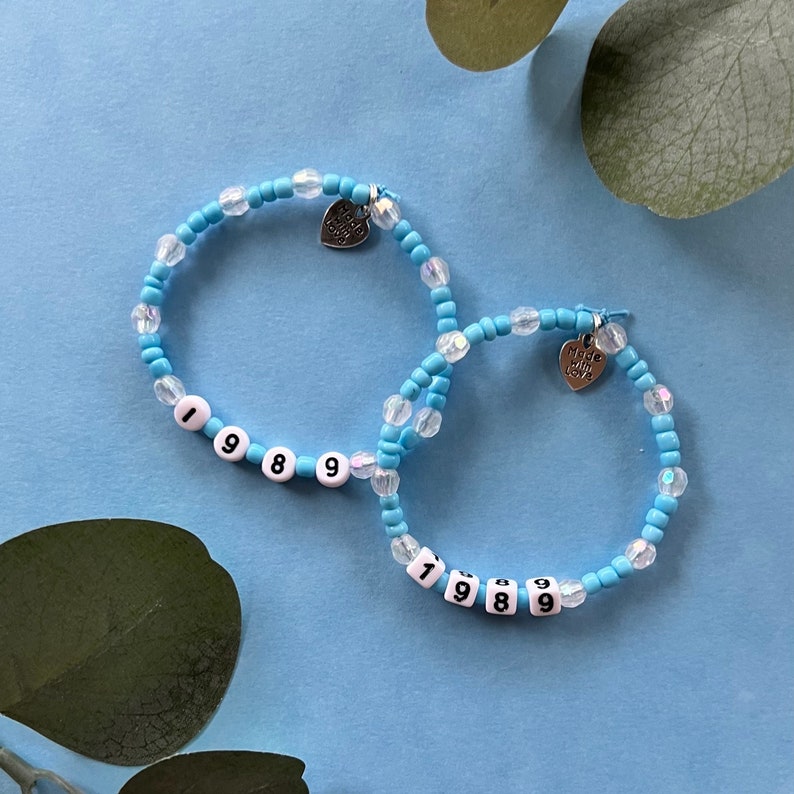 May include: Two beaded bracelets. The bracelets are light blue with clear, iridescent beads and white letter beads spelling "1989". Each bracelet has a small silver heart charm that says "Made with love".