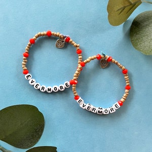May include: Two beaded bracelets with the word "EVERMORE" spelled out in white letter beads. The bracelets feature gold and red beads, and small heart charms that read "Made with Love".