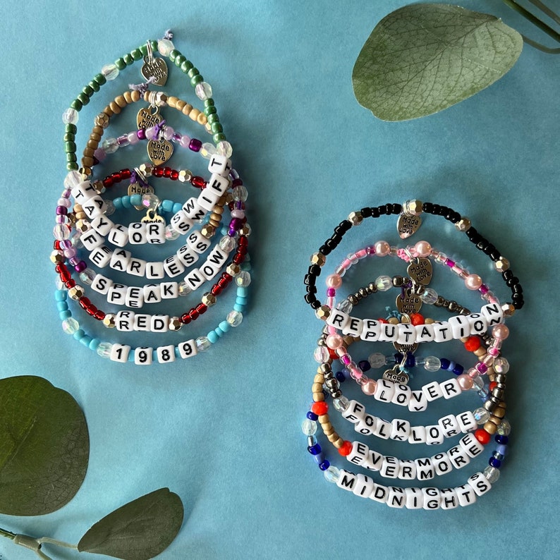 Pode incluir: Uma cole&ccedil;&atilde;o de pulseiras de contas com letras brancas que soletram palavras como "Taylor", "Fearless" e "Reputation". As pulseiras s&atilde;o de v&aacute;rias cores, incluindo vermelho, azul e dourado, e t&ecirc;m pequenos pingentes.