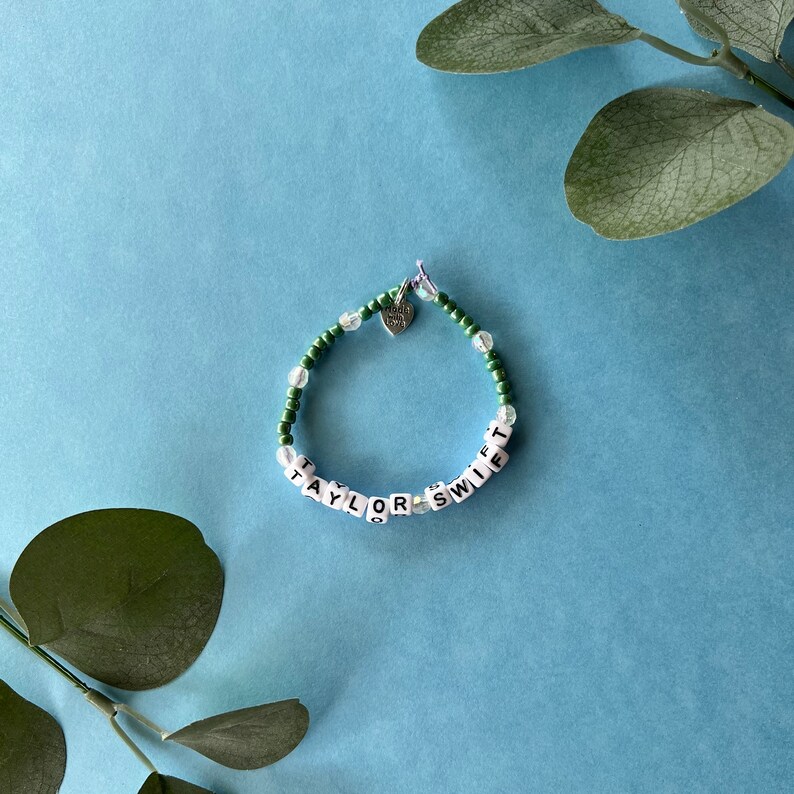 Pode incluir: Uma pulseira de contas com o nome "TAYLOR SWIFT" escrito com contas de letras brancas. A pulseira apresenta contas verdes e transparentes e um pequeno pingente de cora&ccedil;&atilde;o. Fundo azul claro com folhas verdes.