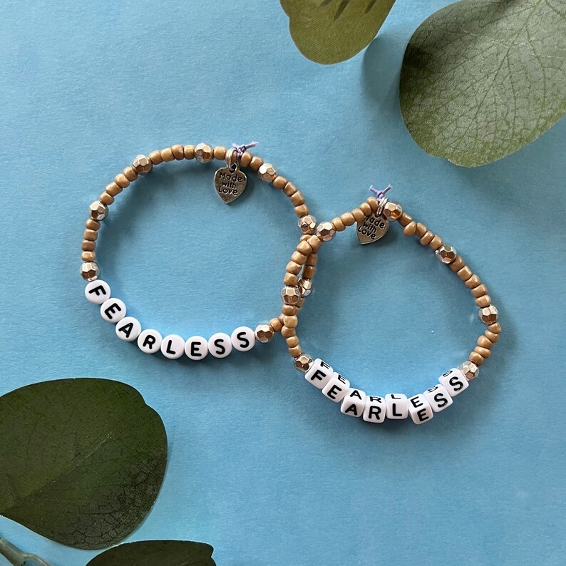 May include: Two beaded bracelets with the word "FEARLESS" spelled out in white letter beads. The bracelets have gold and silver beads and small heart charms that say "Made with Love". The bracelets are on a light blue surface.