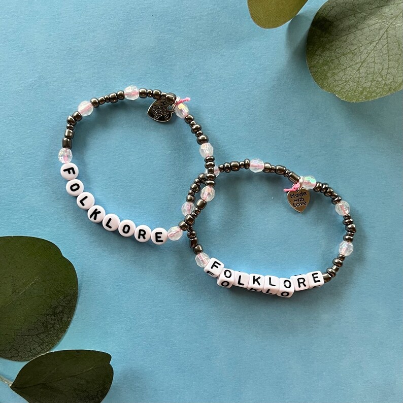 May include: Two beaded bracelets with the word "FOLKLORE" spelled out in white letter beads. The bracelets have a mix of dark gray and clear beads, with small heart charms that say "Made with Love".