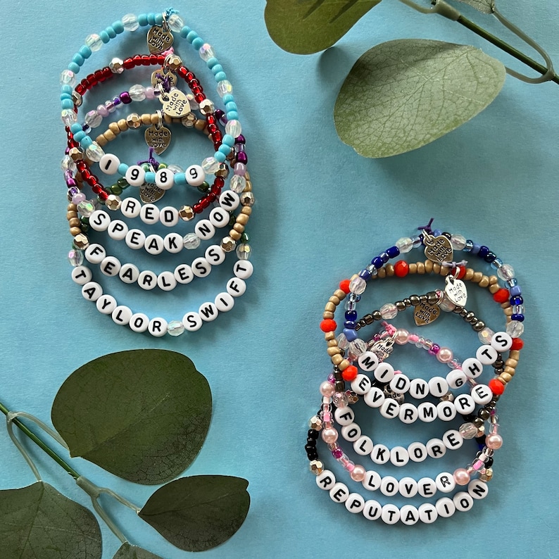 May include: Several beaded bracelets with white letter beads spelling out song titles and the name "Taylor Swift". The bracelets feature various colors of beads, including blue, red, gold, and pink. Each bracelet has a small heart charm.