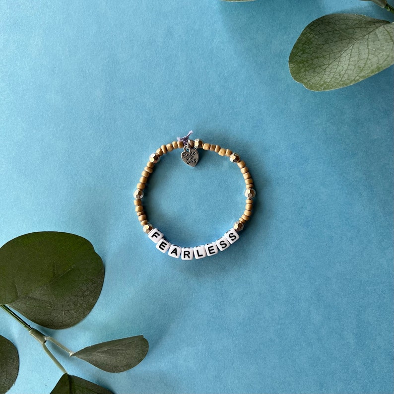 Pode incluir: Uma pulseira de contas com a palavra "FEARLESS" escrita em contas de letras brancas. A pulseira apresenta uma mistura de contas douradas e prateadas, e um pequeno pingente de cora&ccedil;&atilde;o que diz "Made with Love". A pulseira est&aacute; em um fundo azul.