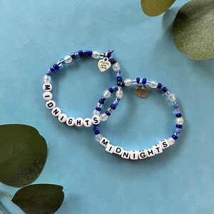 May include: Two beaded bracelets with the word "MIDNIGHTS" spelled out in white letter beads. The bracelets feature blue and clear beads and small heart charms that say "Made with Love".