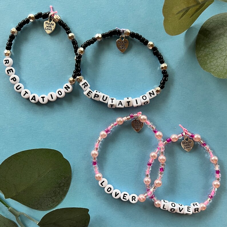 May include: Four beaded bracelets. Two black bracelets spell "REPUTATION" with white letter beads. Two pink bracelets spell "LOVER" with white letter beads. Each bracelet has a heart charm that says "Made with Love."