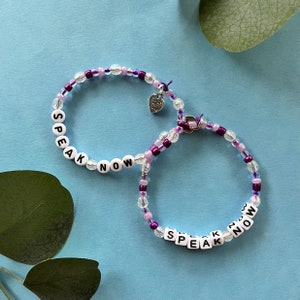 May include: Two beaded bracelets with the words "SPEAK NOW" spelled out in white letter beads. The bracelets feature clear, purple, and lavender beads. A small heart charm with the words "made with love" hangs from one bracelet. The background is light blue.