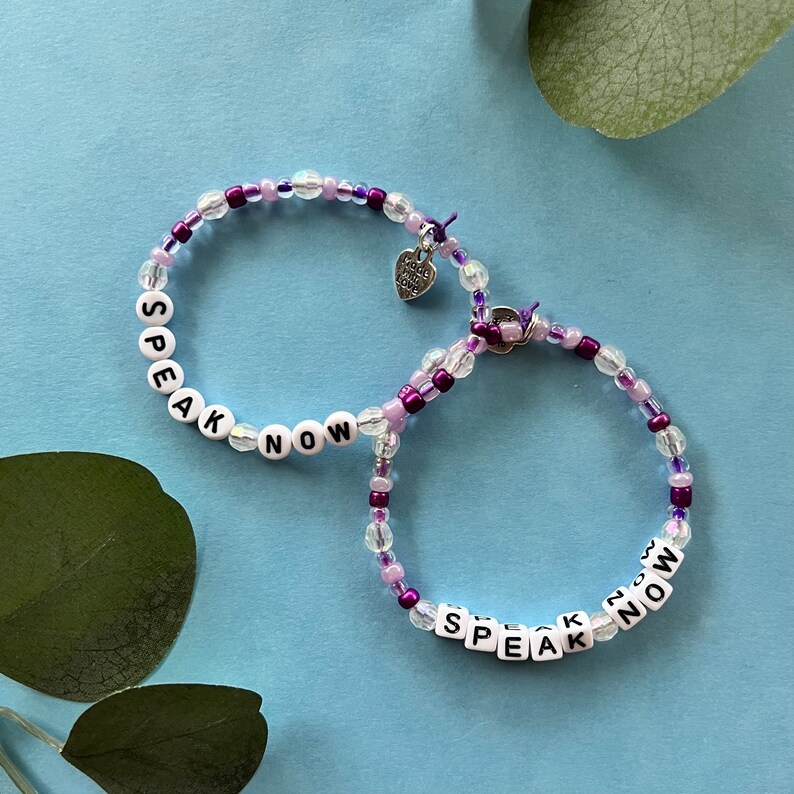 May include: Two beaded bracelets with the words "SPEAK NOW" spelled out in white letter beads. The bracelets feature clear, purple, and lavender beads. A small heart charm with the words "made with love" hangs from one bracelet. The background is light blue.