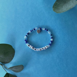 Pode incluir: Uma pulseira de contas azuis e transparentes com a palavra "MIDNIGHTS" escrita em contas de letras brancas. Um pequeno pingente de cora&ccedil;&atilde;o prateado com a inscri&ccedil;&atilde;o "Made with Love" pende da pulseira.