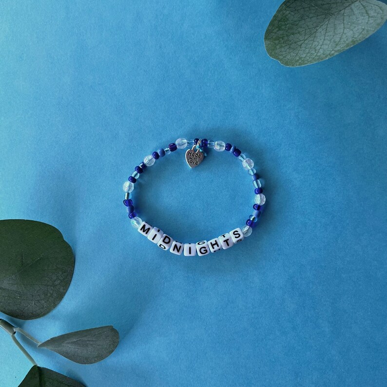 Pode incluir: Uma pulseira de contas azuis e transparentes com a palavra "MIDNIGHTS" escrita em contas de letras brancas. Um pequeno pingente de cora&ccedil;&atilde;o prateado com a inscri&ccedil;&atilde;o "Made with Love" pende da pulseira.